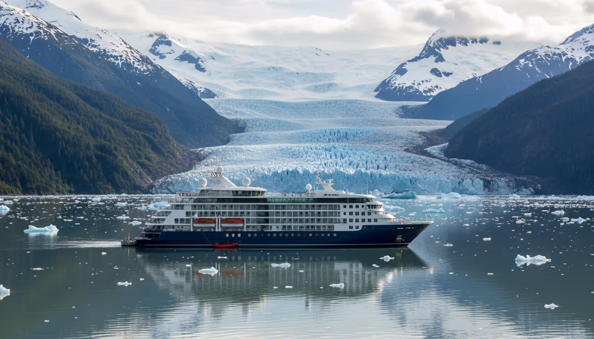 Luxury small cruise ship anchored in front of a massive blue tidewater glacier in an Alaskan fjord surrounded by snow-capped mountains