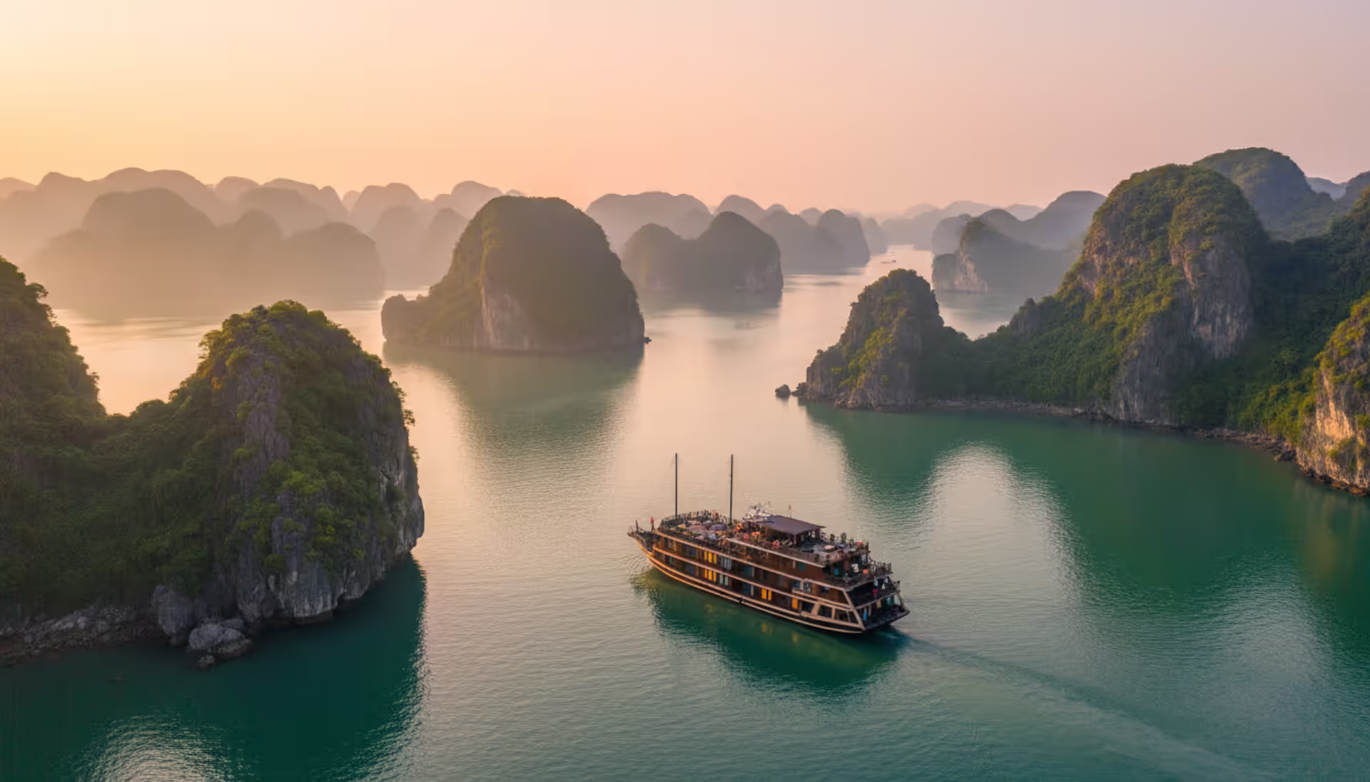 Luxury wooden cruise ship sailing through Halong Bay at sunrise with limestone karst formations rising from emerald water and morning mist