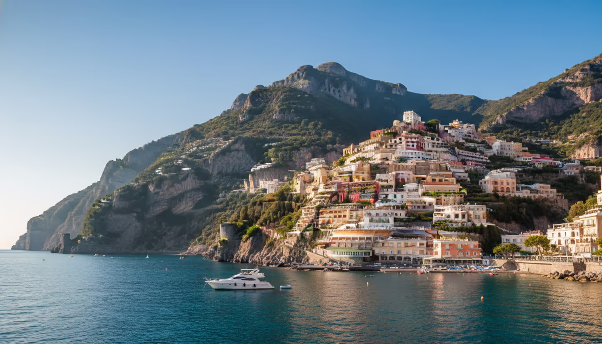 Panoramic view of the Amalfi Coast from the sea with colorful cliffside villages, turquoise water, and a white yacht anchored in a cove